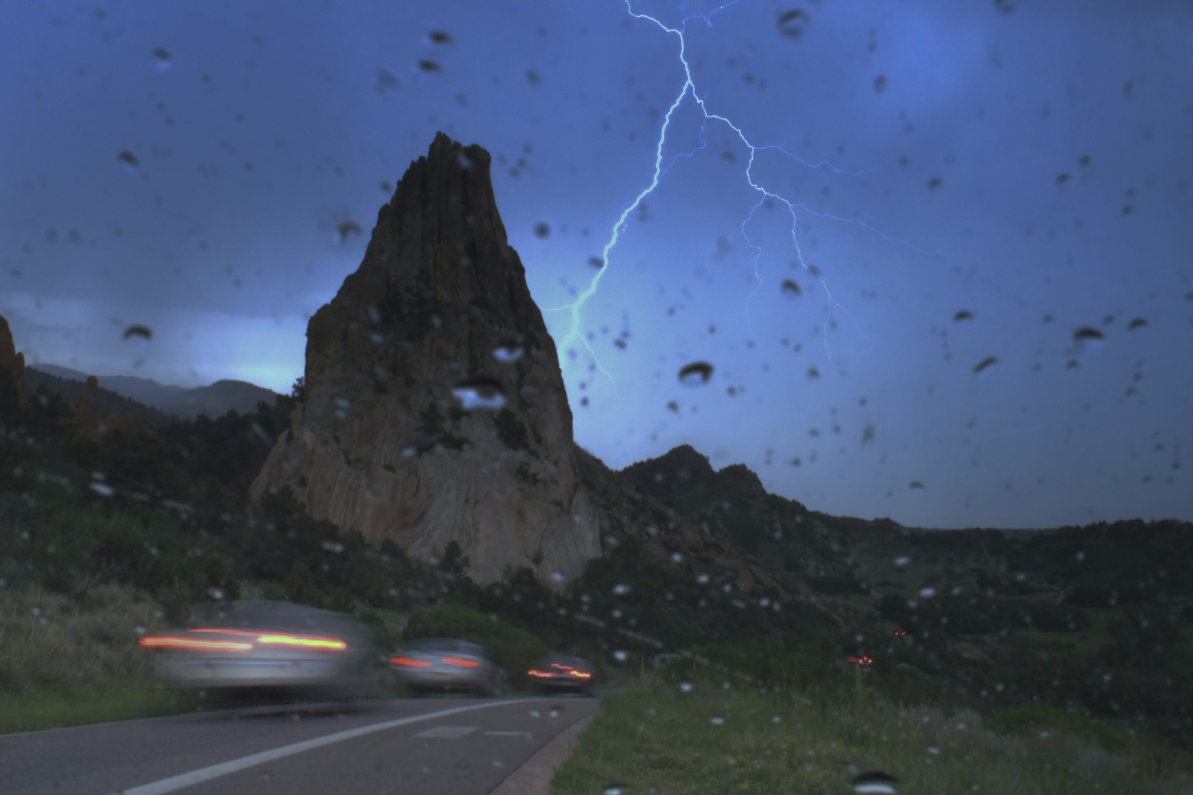 Lightning strike in sky on a mountain road with a rainy windshield and car lights.